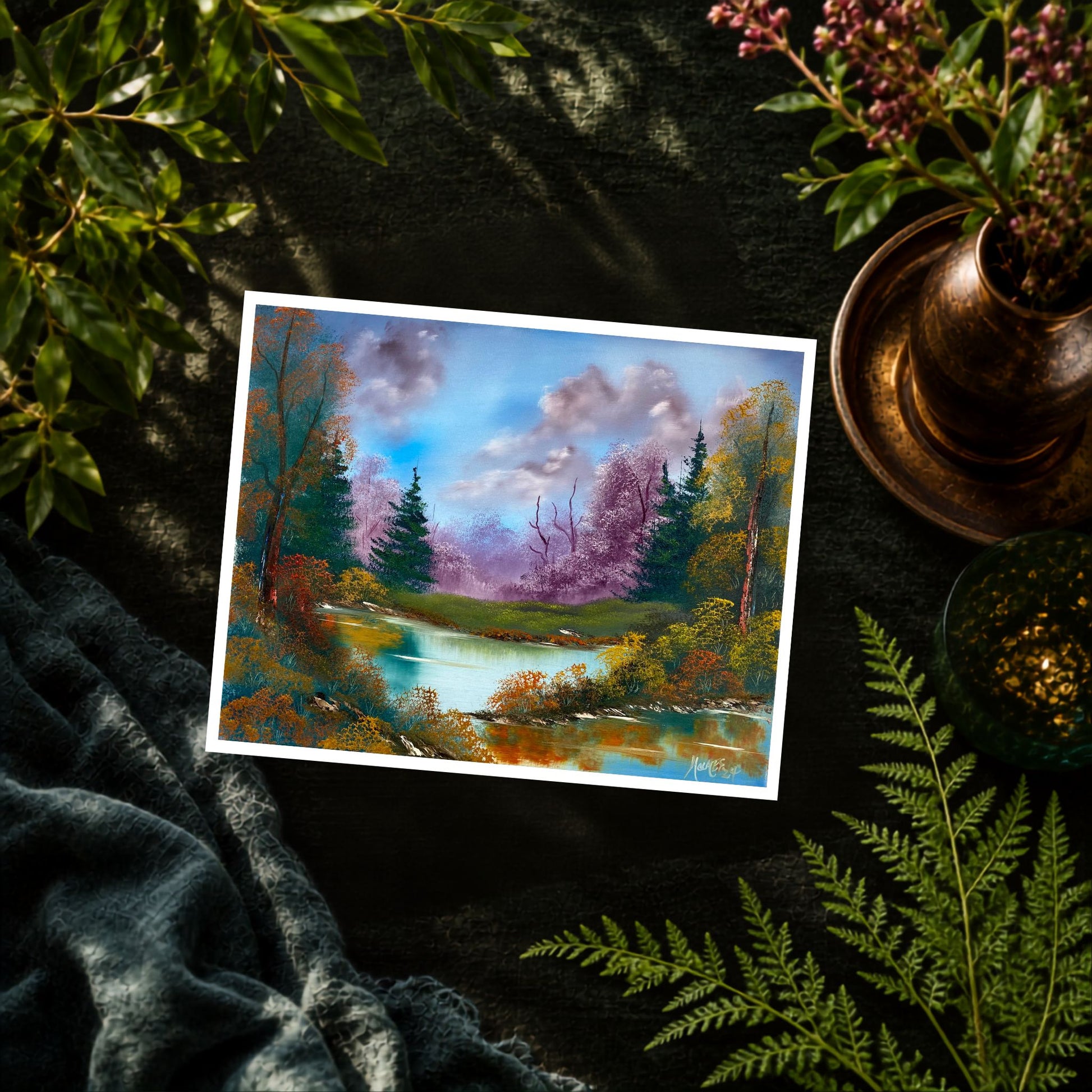 A landscape art print depicting a colorful river scene with trees and a blue sky, styled on a dark textured surface with green leaves, a blue blanket, and a bronze-colored dish.