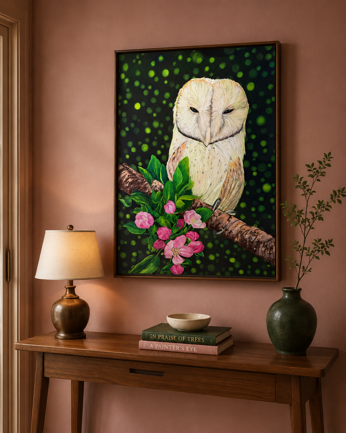 A framed original oil painting of a barn owl perched on a branch with pink flowers, displayed on a dusty rose wall above a wooden console table with a lamp, books, and a vase.