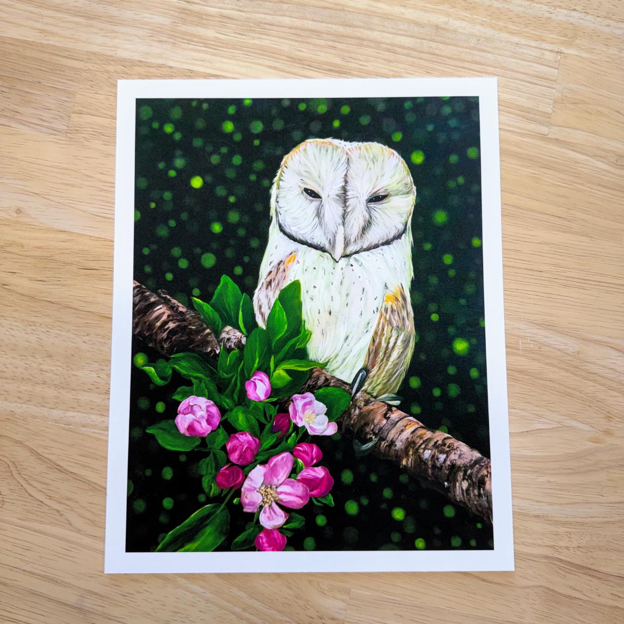 A rectangle art print featuring a detailed illustration of a barn owl perched on a branch with pink flowers, set against a dark green bokeh background, displayed on a light wood surface.