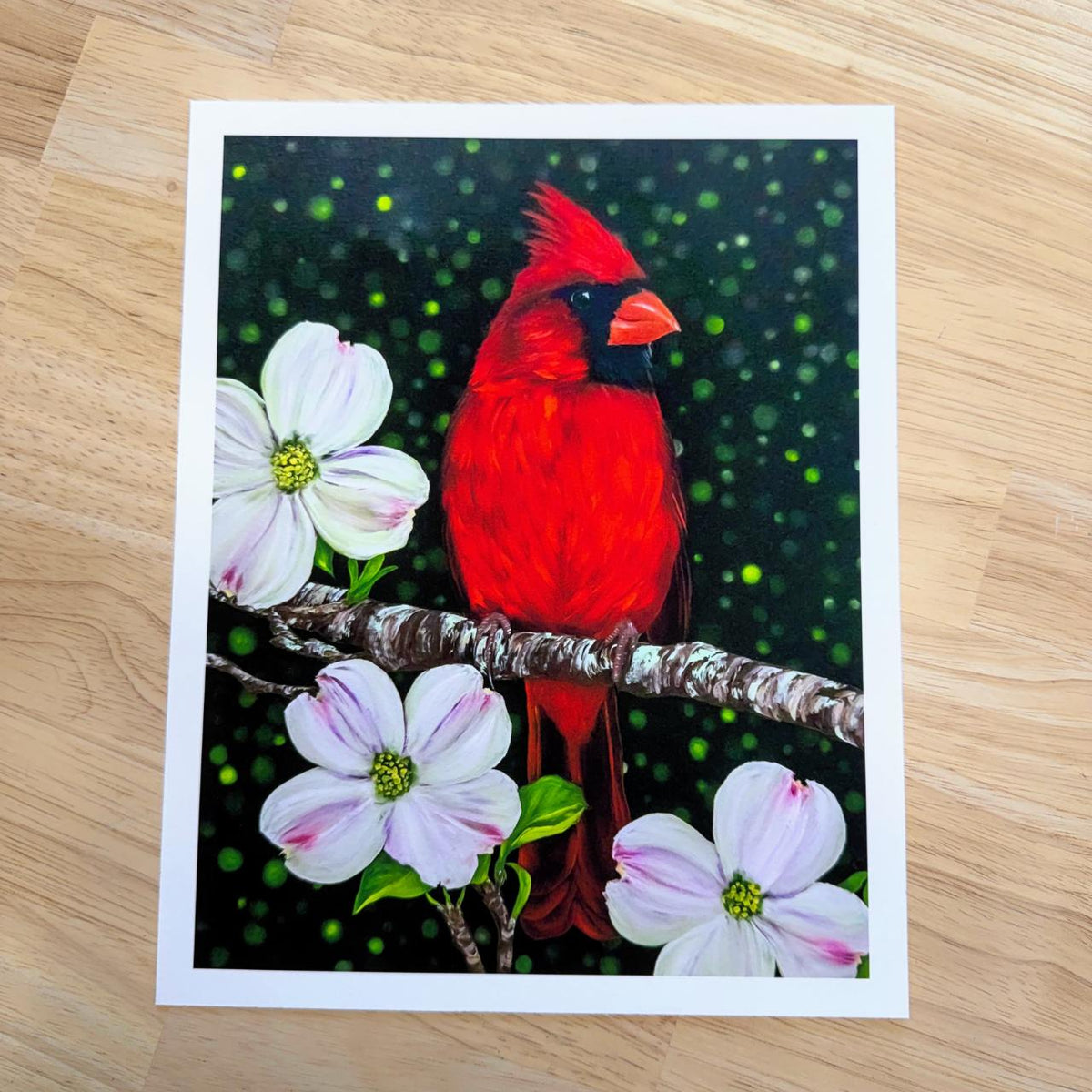 A rectangle art print featuring a bright red cardinal perched on a branch with white and pink dogwood flowers against a dark green bokeh background, displayed on a light wood surface.