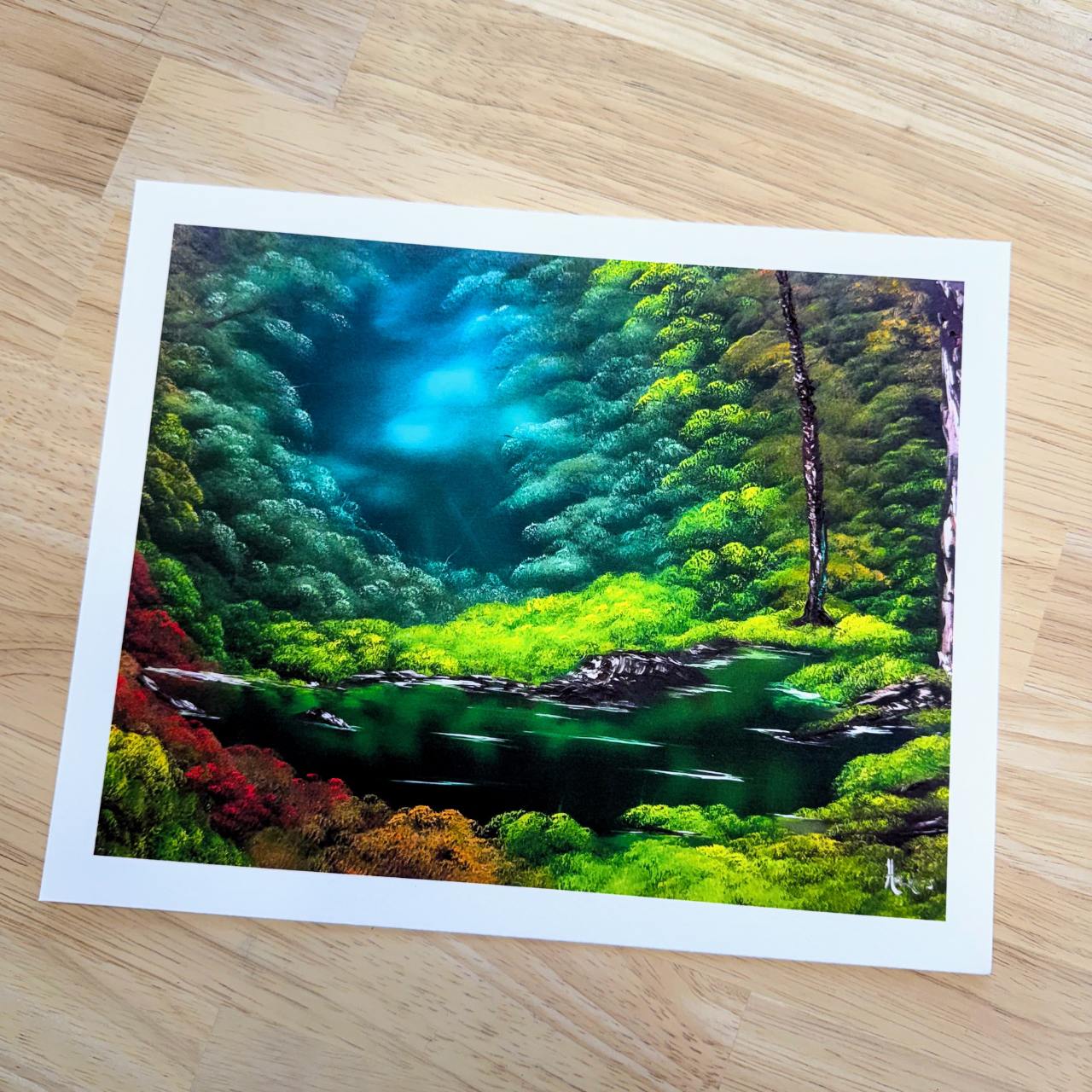 A rectangle art print depicting a vibrant, colorful forest landscape with a river, shown on a light wood floor. The print features a white border and is signed by the artist in the bottom right corner.
