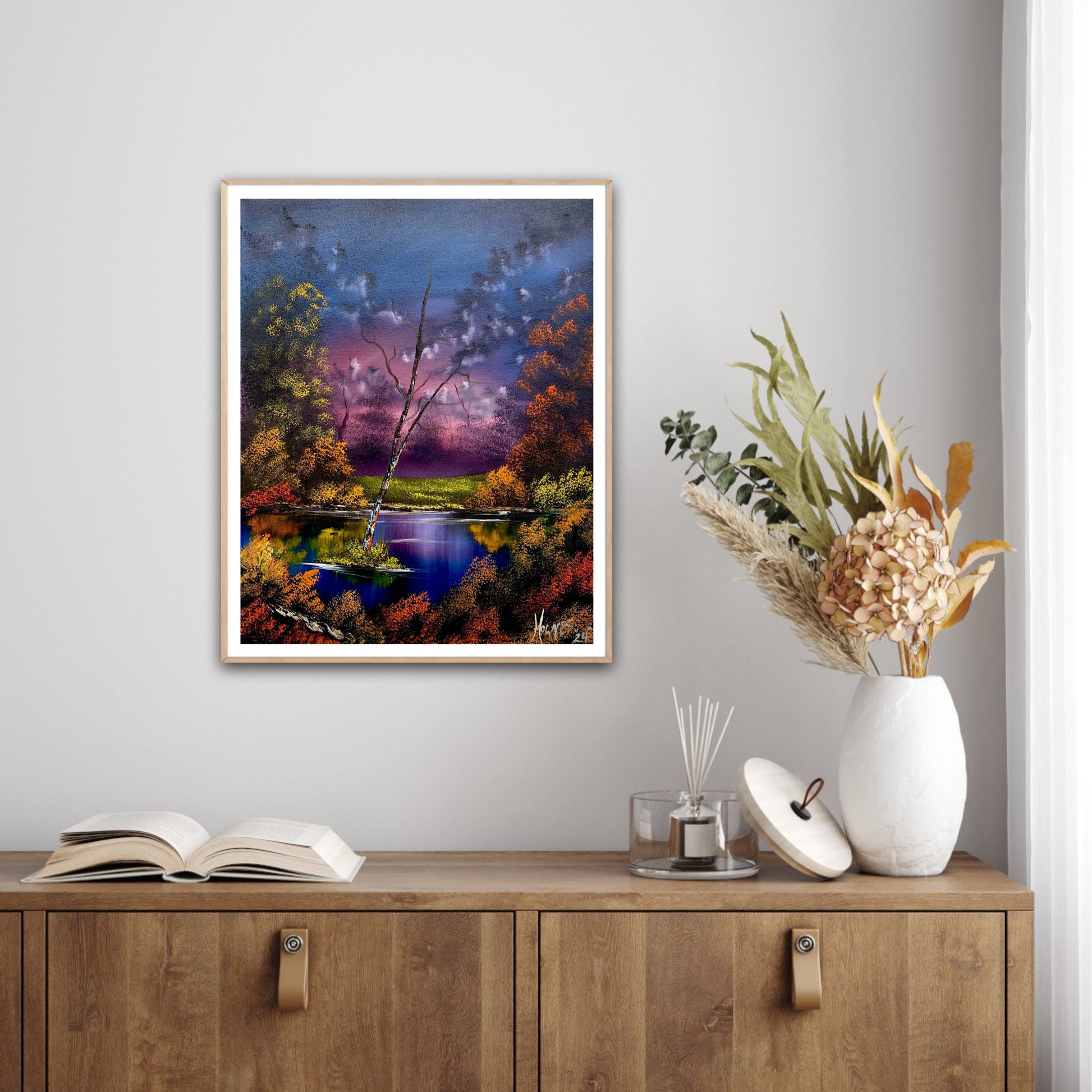 Framed artwork of a landscape with a river and trees on a wall above a wooden cabinet.