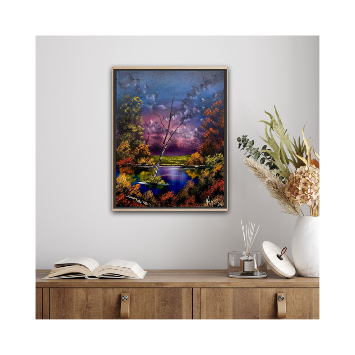 Framed painting of a landscape with a river and trees on a wall above a wooden cabinet.