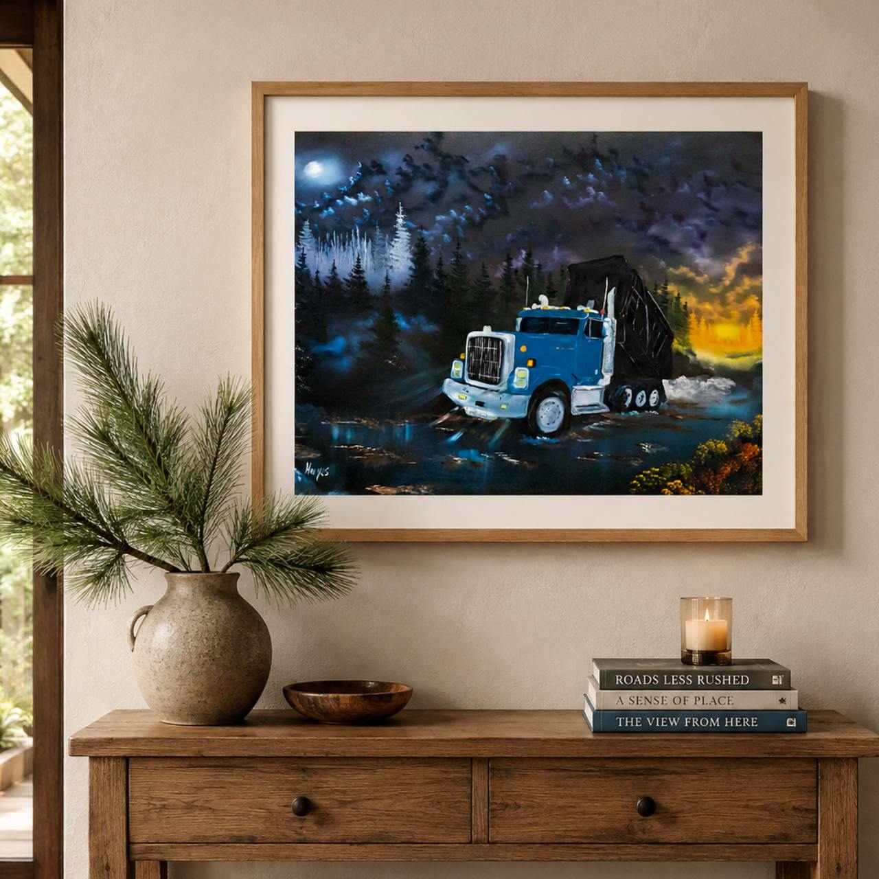 A framed art print depicting a blue semi-truck drivingon a gravel road at night under a full moon, displayed on a cream wall above a rustic wooden console table with a vase of pine branches and a candle.