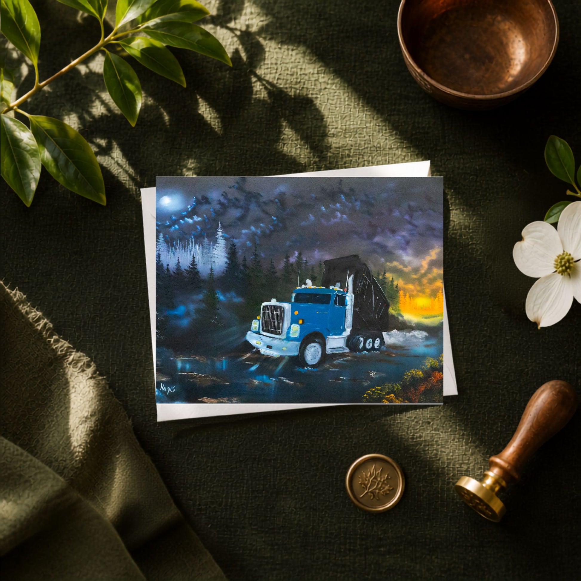 A greeting card showing a blue dump truck driving through water at dusk, styled on a dark green fabric surface with leaves, a copper bowl, a white flower, and a wooden stamp.