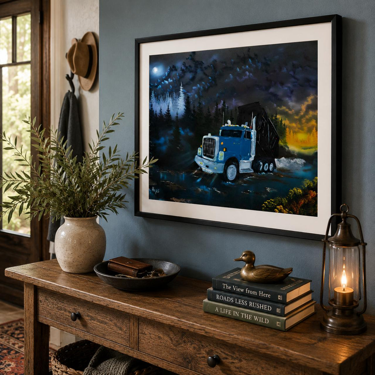 A framed art print depicting a blue semi-truck driving through a dark, moonlit forest road at night, styled on a rustic wooden console table in a cozy room setting with a lantern, duck figurine, and books.