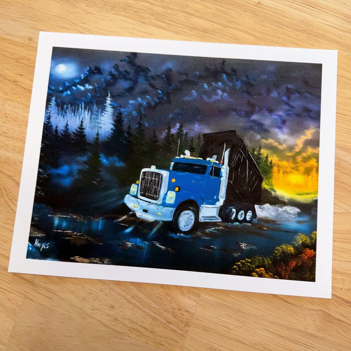 A rectangular art print featuring a blue dump truck driving through a gravel road at night under a dramatic cloudy sky, placed on a light wood surface.