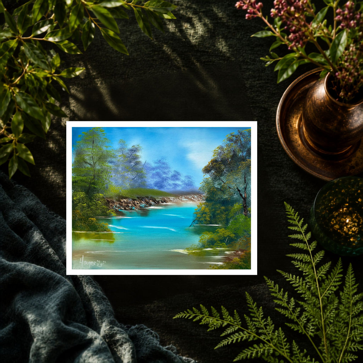 A landscape art print depicting a scenic river with turquoise water, green trees, and a blue sky, styled on a dark textured surface with ferns, a copper pot, and a gray blanket.