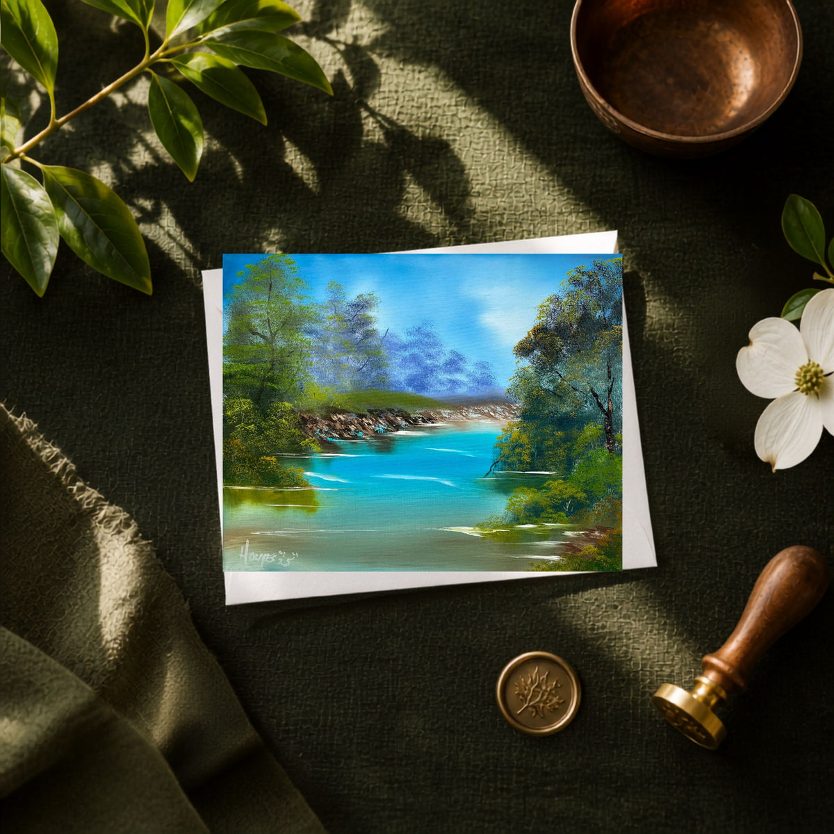 A greeting card showing a colorful landscape painting of a river surrounded by green trees, styled on a dark textured fabric with a copper bowl, white flower, wax seal, and wooden stamp nearby.