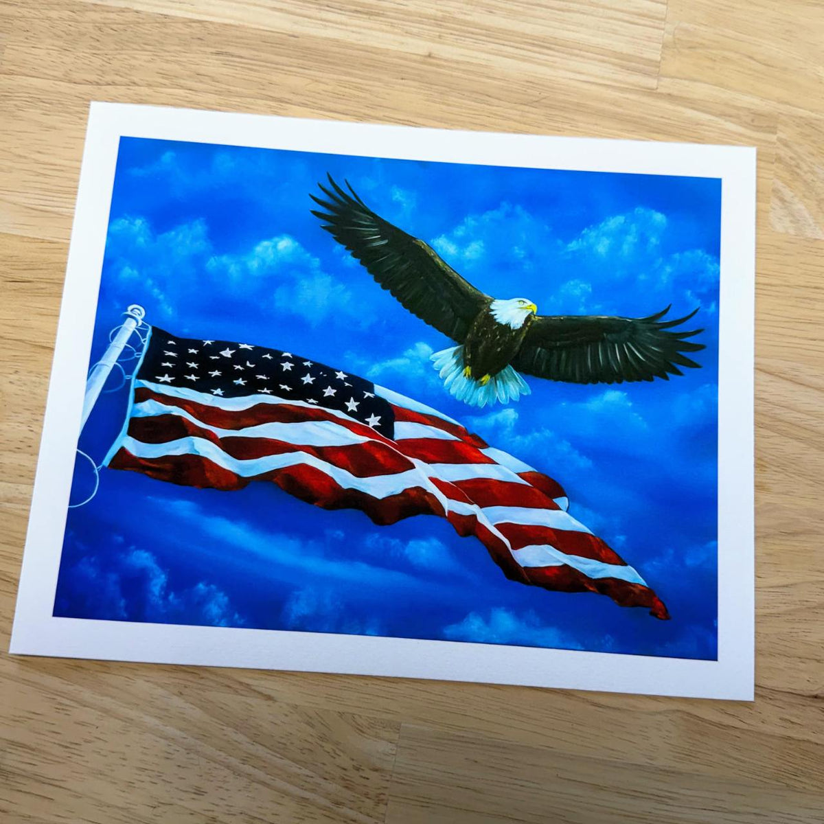 A rectangle art print featuring a bald eagle flying above a waving American flag against a bright blue sky, photographed on a light wood floor.