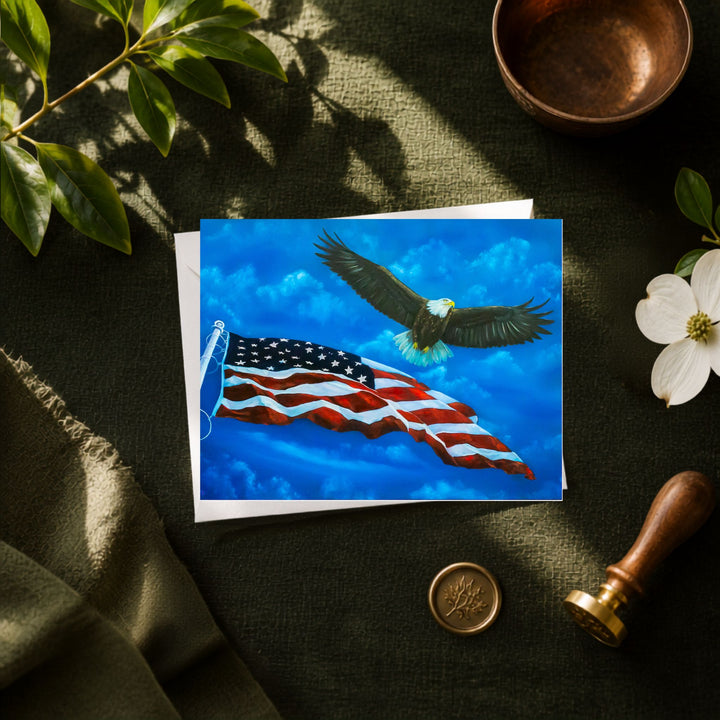 A greeting card of a bald eagle flying over a waving American flag, styled in a flat-lay on a dark green textured fabric with a copper bowl, white flower, green leaves, and a wax seal stamp.