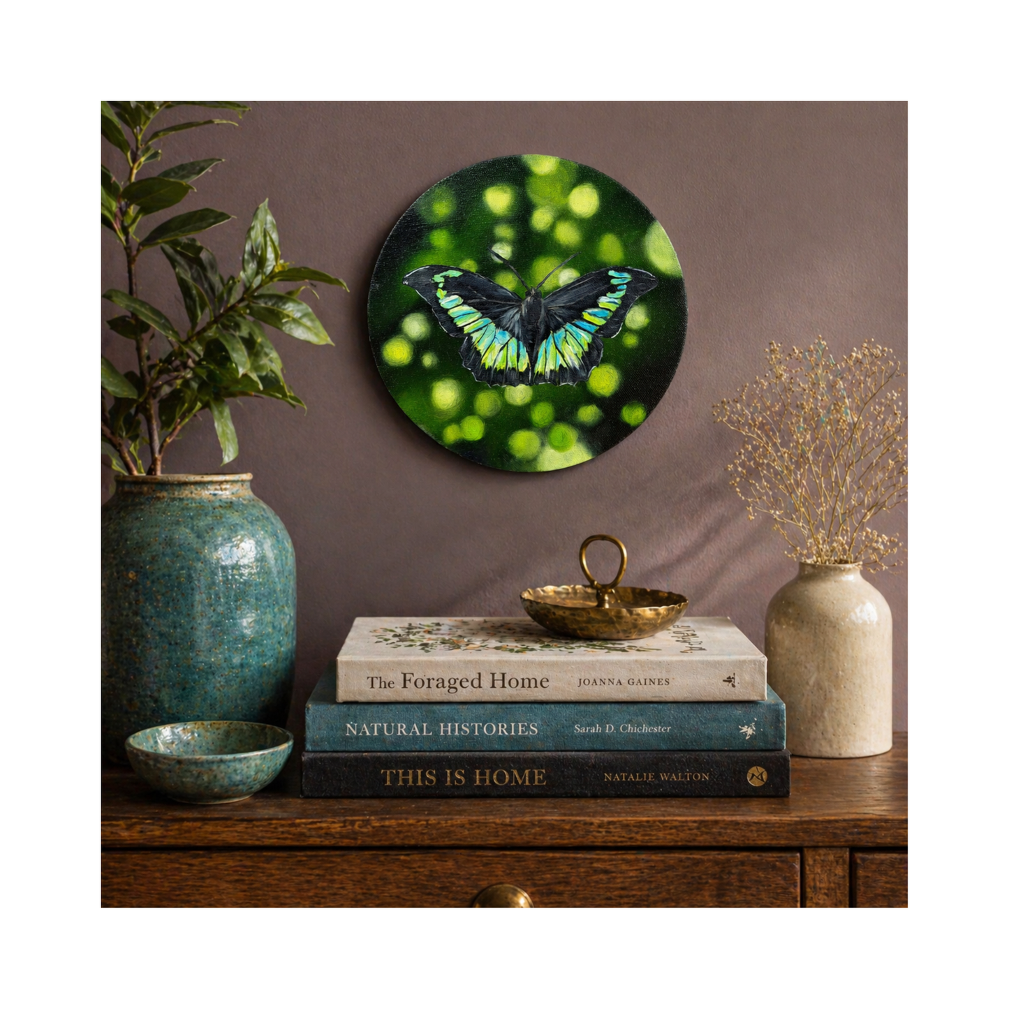 Teal green buttefly oil painting on a round canvas on a wall above books and vases.