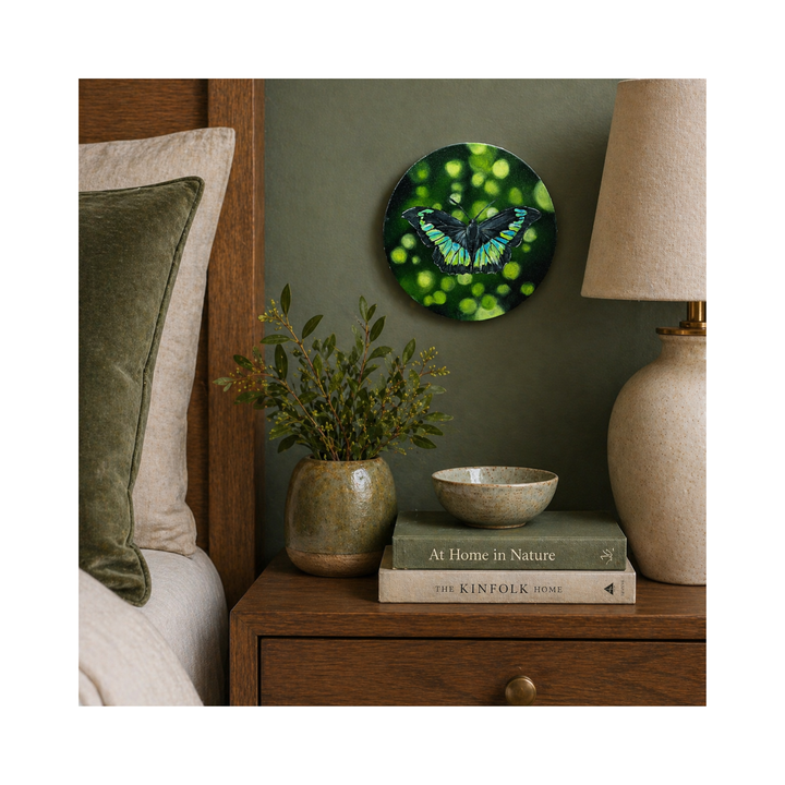 Round canvas oil painting with a blue green butterfly on a wall above a wooden nightstand with books and a lamp.