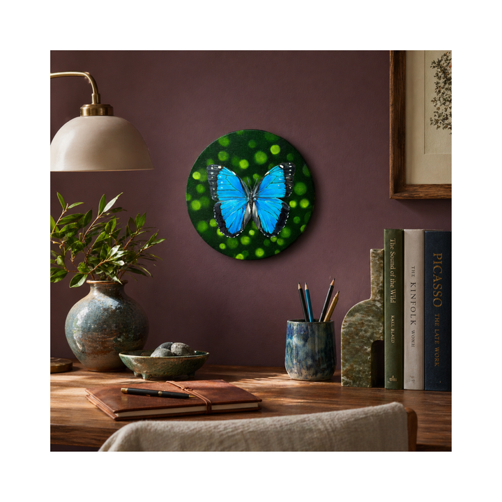 Morho butterfly oil painting on round canvas on a plum wall over desk with books, flower vase and a journal