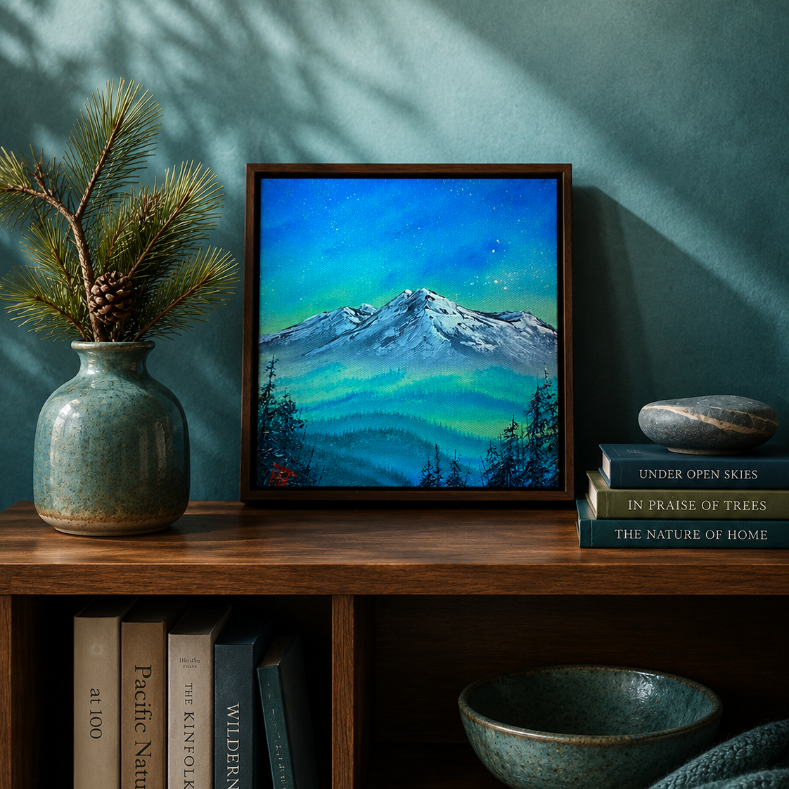 A framed landscape painting of a snow-capped mountain under a vibrant blue and green night sky, styled on a wooden shelf next to a ceramic vase with pine branches and a stack of books.