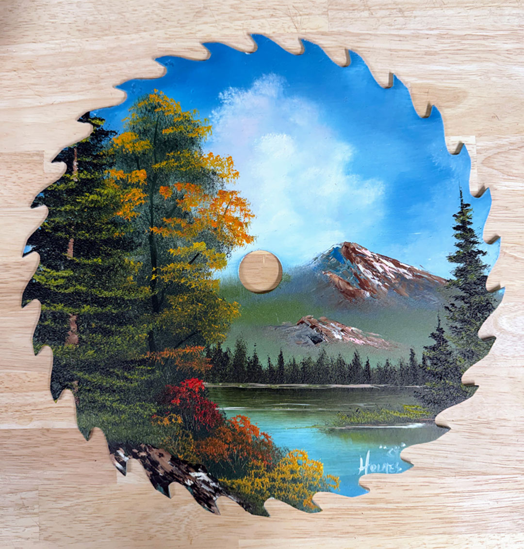 A circular saw blade with a painted landscape design, featuring a blue sky, white clouds, green and yellow trees, a lake, and mountains, displayed on a light wood background.