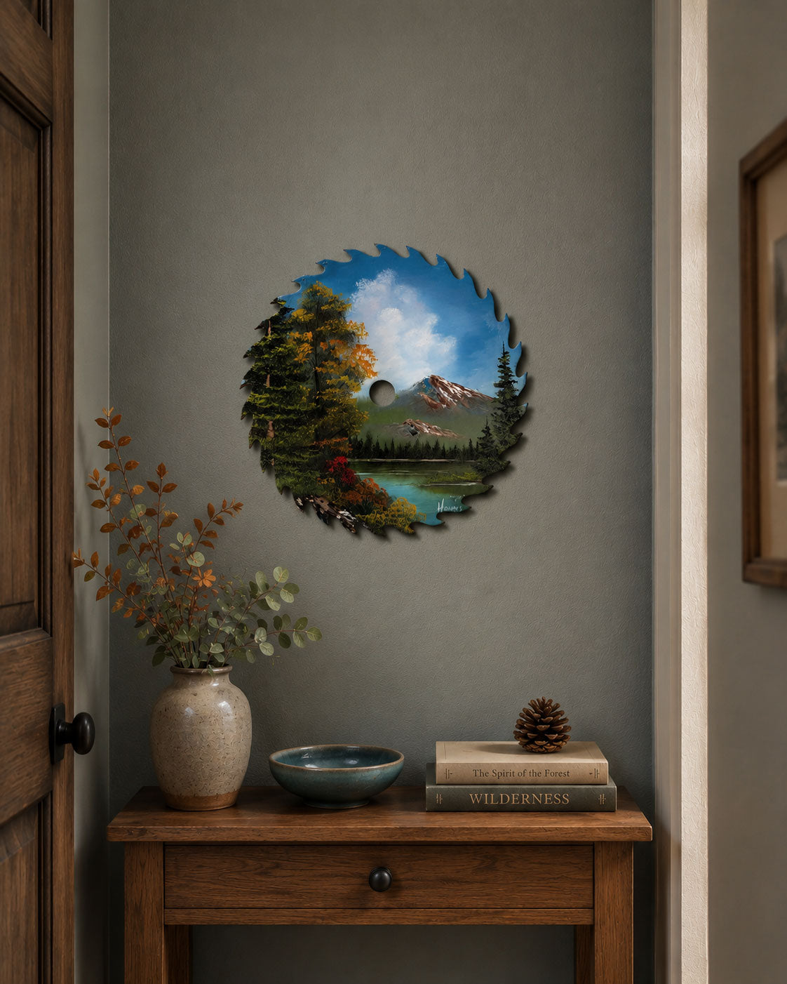 A circular wall art piece featuring a scenic mountain lake landscape with evergreen trees and a blue sky, mounted on a gray wall above a wooden console table with a vase, bowl, and books.