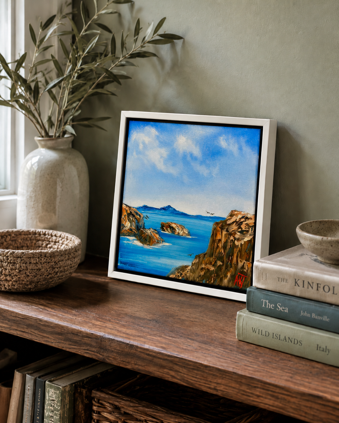 Framed artwork of a coastal scene on a wooden shelf with books and a vase.