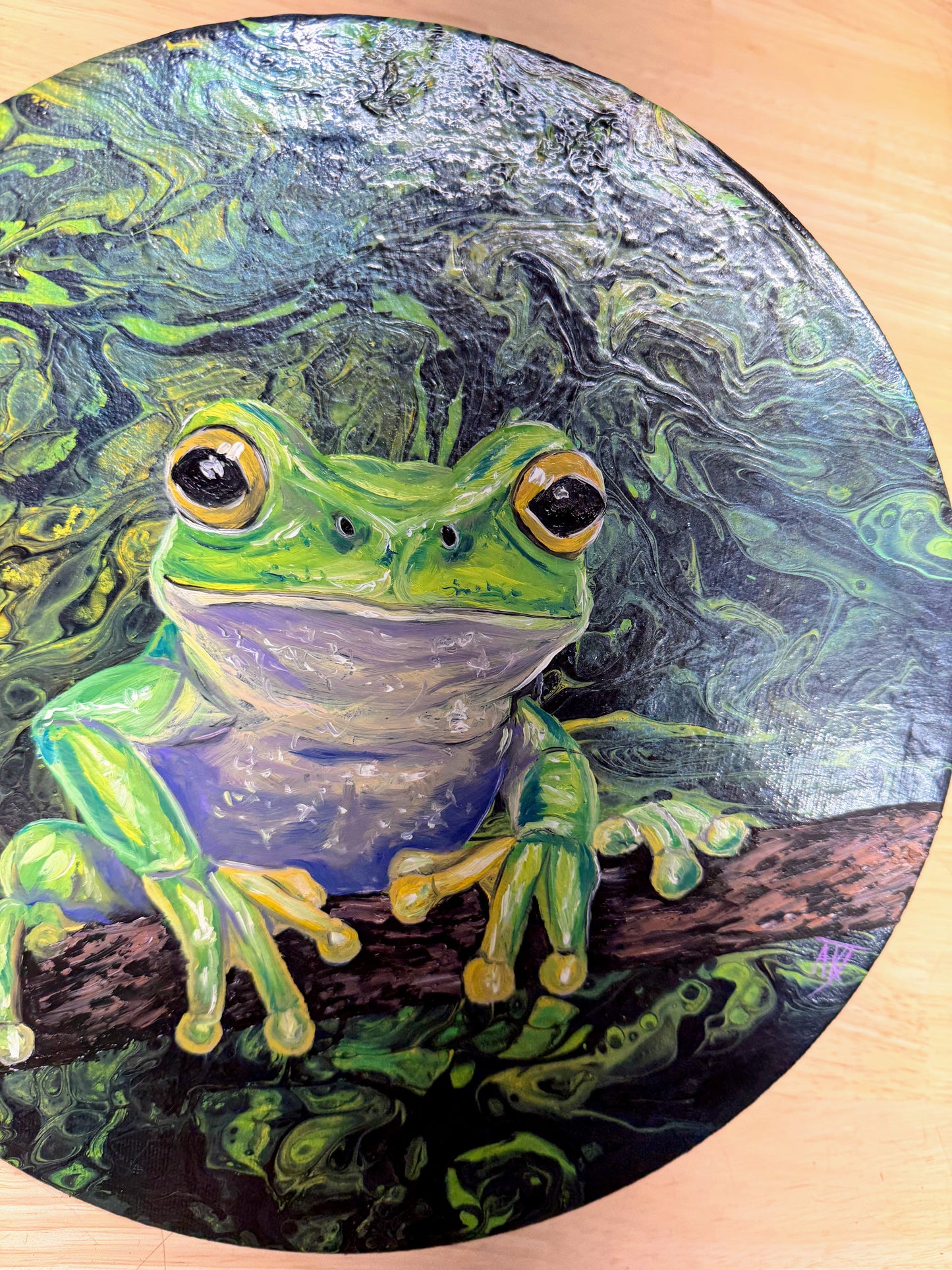 A circular painted artwork of a bright green tree frog perched on a branch, with swirling green, blue, and black background textures. The painting is photographed on a light wood surface.