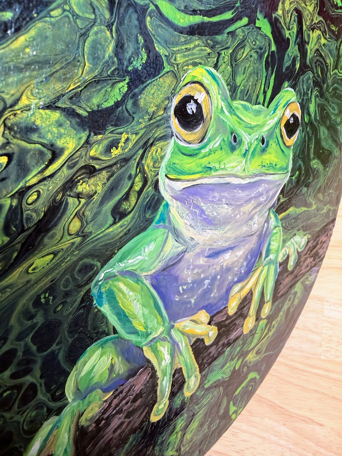 A close-up of a painted green tree frog perched on a branch, with swirling dark green and black background details and a light wood surface visible at the lower right.