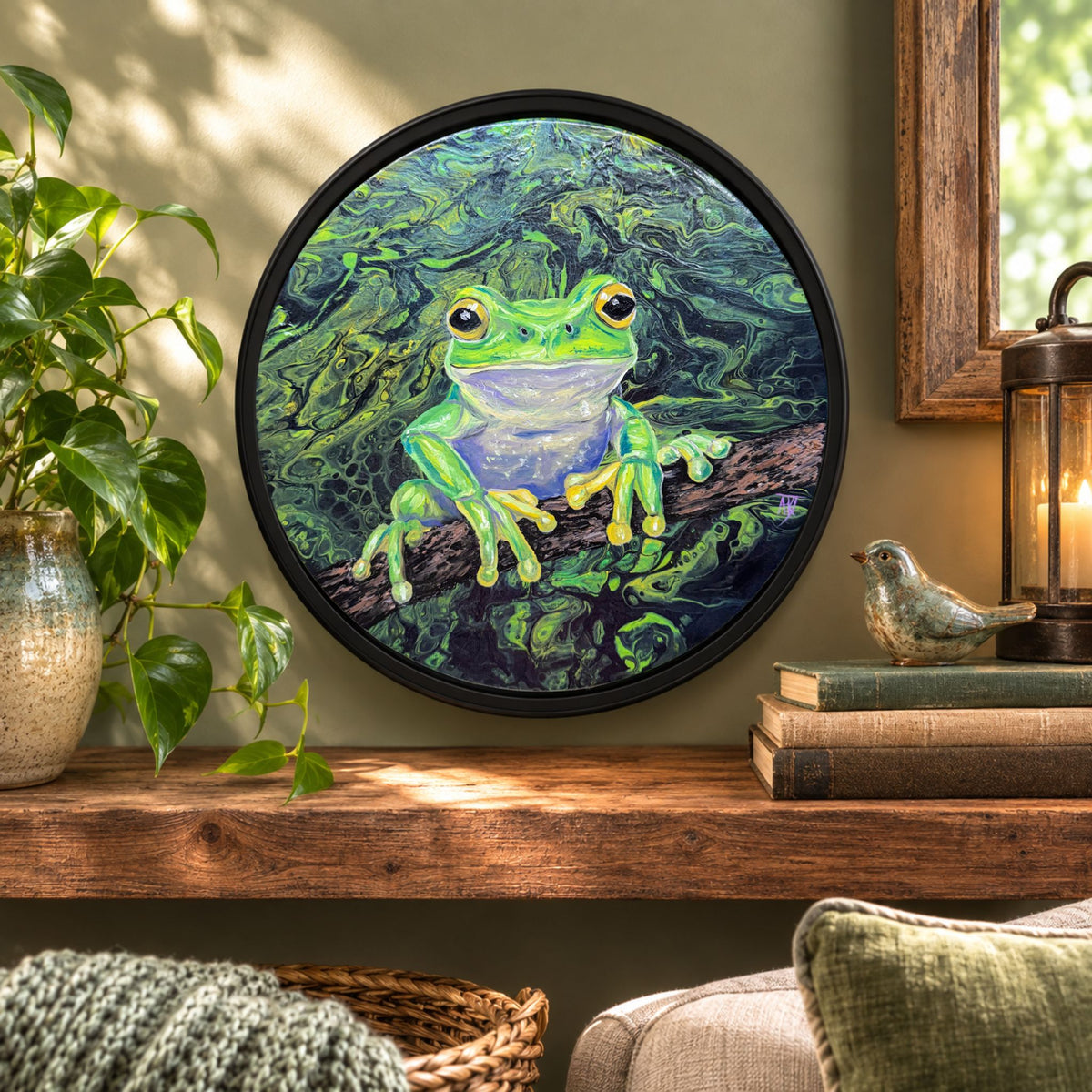 Round framed wall art featuring a bright green frog perched on a branch against a soft sage and green leafy background, displayed on a wooden shelf in a styled room with a plant, books, and a lantern.