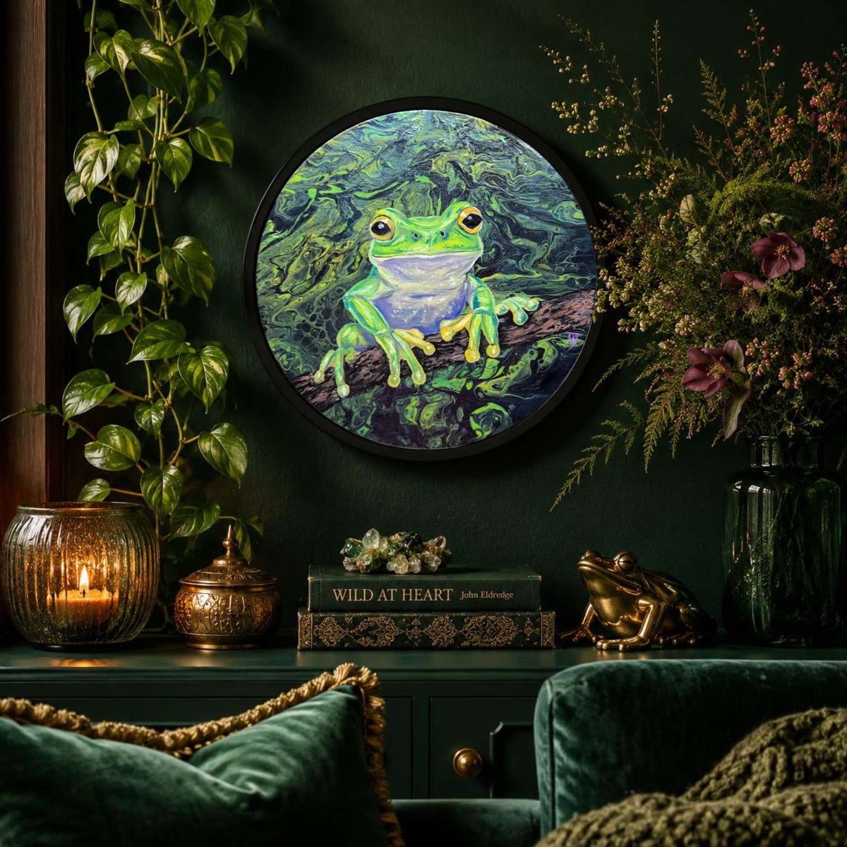A round framed wall art featuring a bright green tree frog on a dark forest background, displayed in a styled room setting with a dark green velvet sofa, plants, and decorative objects.