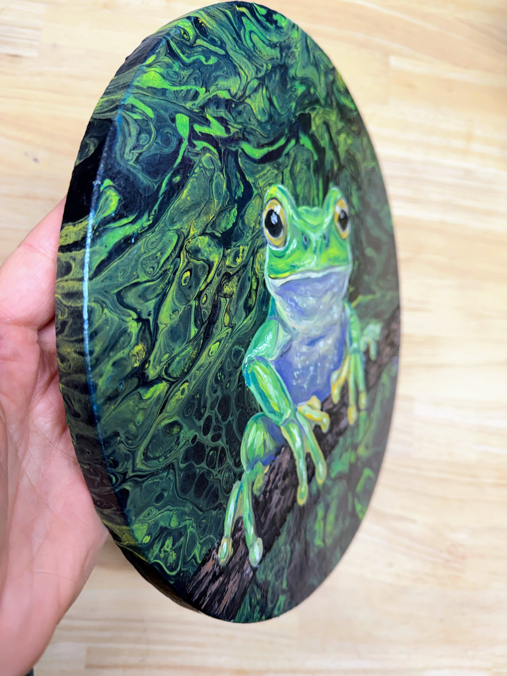 A hand holds a round canvas painting featuring a green tree frog perched on a branch against a dark, marbled forest background, with a light wood floor behind it.