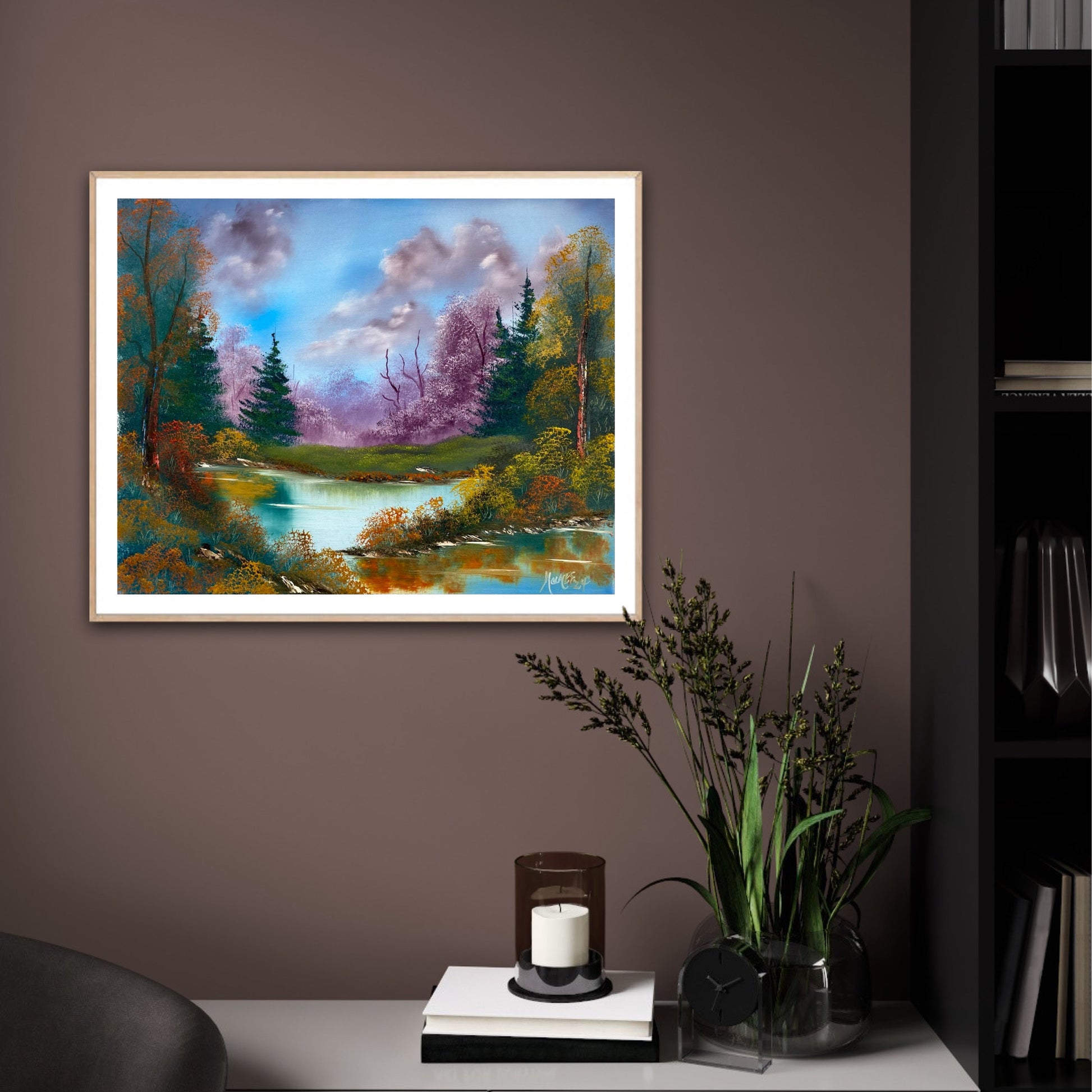 Framed colorful autumn landscape art print on a wall above a cabinet with books and a candle.