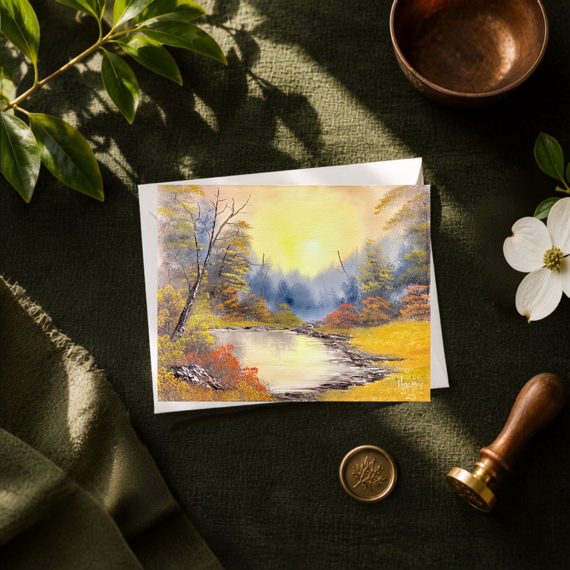 A greeting card of a pond during golden hour displayed on green background with brass bowl and wax seal