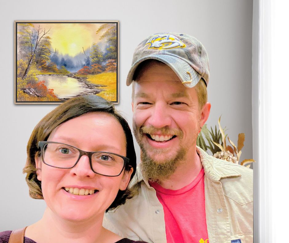 Chris and Anna Holmes, husband-and-wife landscape artists and Certified Bob Ross Instructors, standing together with one of their landscape paintings displayed on the wall behind them.