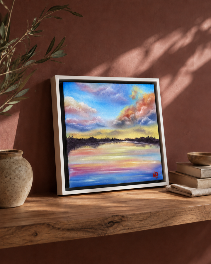 Framed artwork of a sunset over water on a wooden surface with a plant and books in the background.