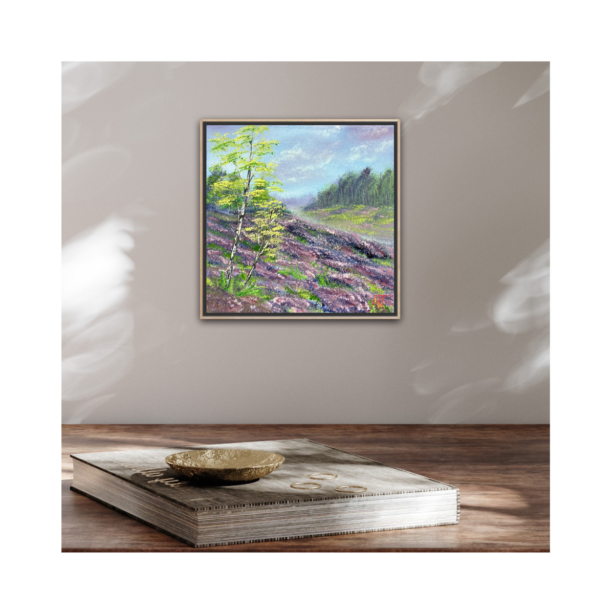 Framed painting of a landscape with trees and heather field on a wall above a wooden table.