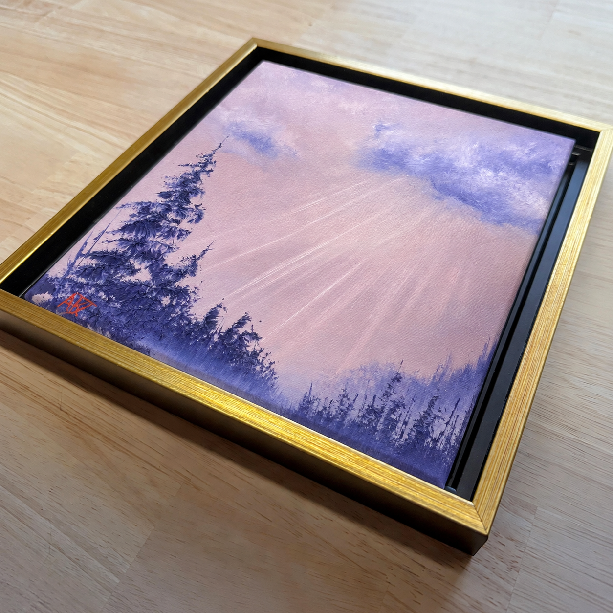 Framed artwork of a landscape with trees and a dusty pink sky laying on a wooden surface