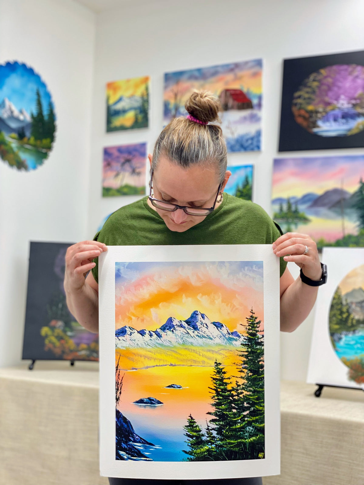 Person holding a framed painting of a mountain landscape in an art gallery.