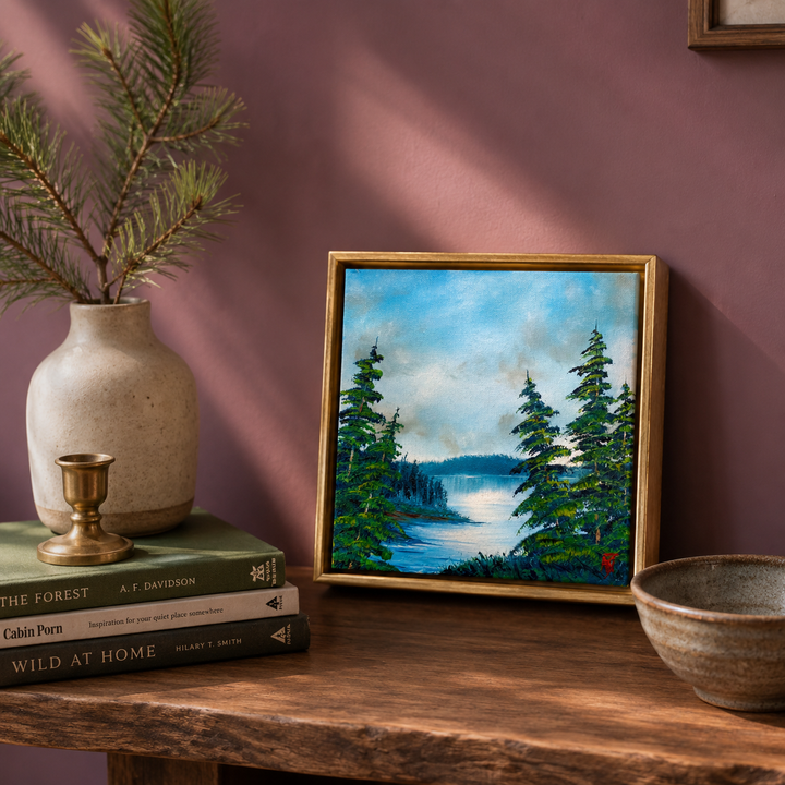 Framed oil painting of a forest and lake scene on a wooden shelf with books and a vase.